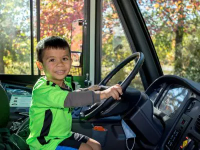 Young boy sitting in drivers seat of an FCPS Electric Bus with his hands on the wheel looking at camera and smiling.