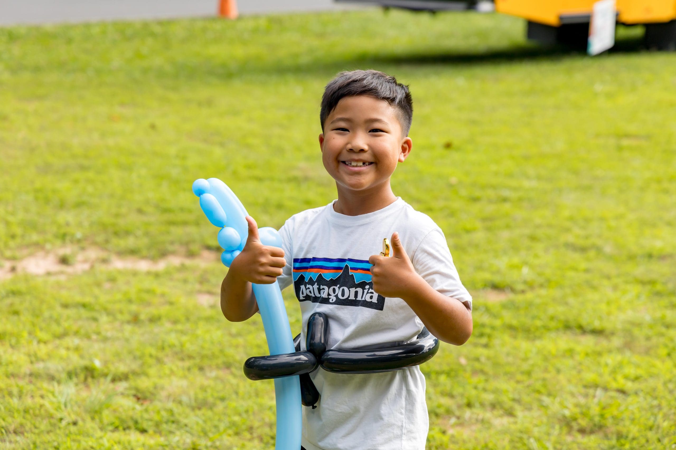Boy standing in grass holding a balloon twisted sword holding two thumbs up and smiling at camera.
