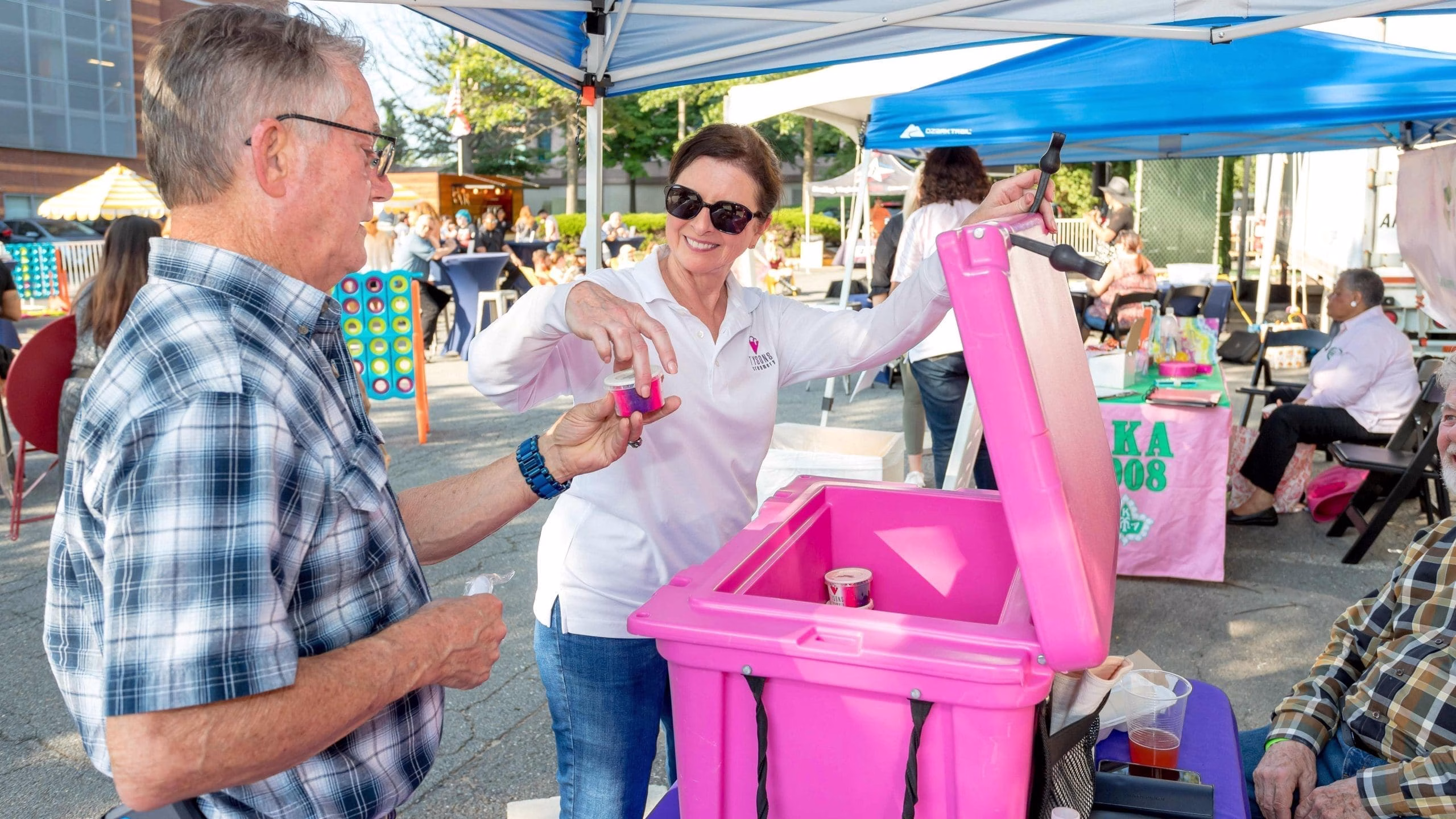 A group of people gathered at an outdoor event, with a pink cooler and a blue tent in the foreground.