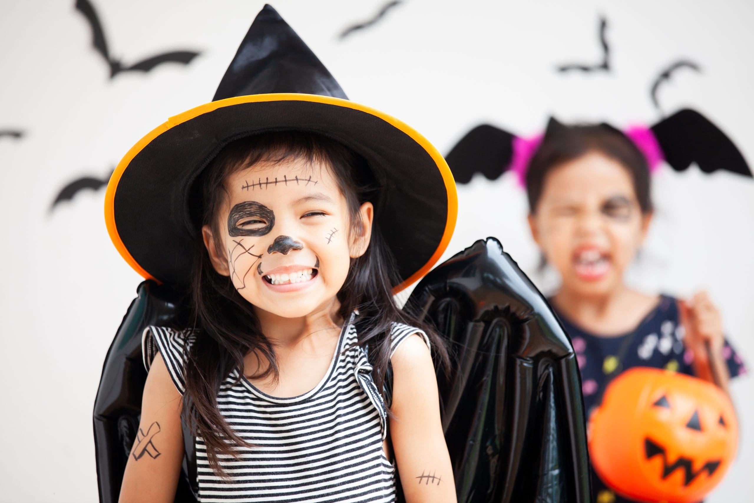 Young girl dressed up for Halloween with big black pointed hat and face painted, smiling at camera.
