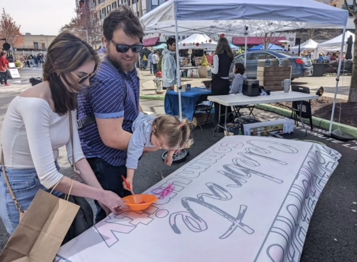 A family of four enjoys a day out at an outdoor market, with a large banner and pizza boxes in the foreground.