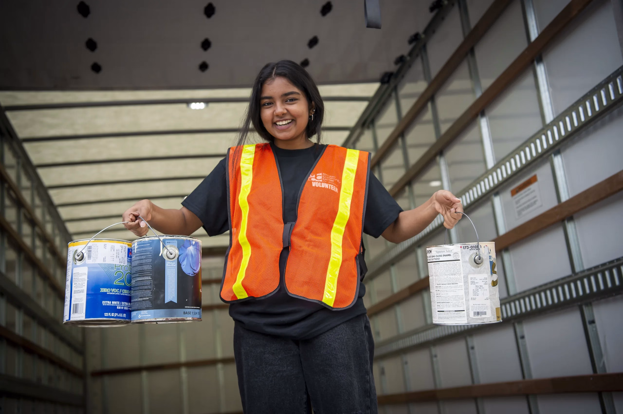 A volunteer in a safety vest holds paint cans in a warehouse, showcasing their dedication to the project.