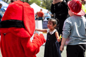A young girl reaches out to touch the hand of a mascot at an outdoor event, surrounded by other children and adults.