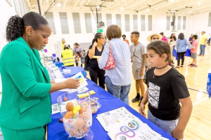 A woman in a green suit standing behind a table with a large glass bowl of candy, talking to a young girl in a black t-shirt