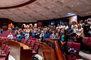 A diverse group of people standing in a large room with red chairs, holding their hands over their hearts.