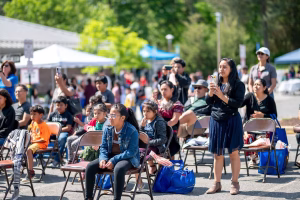 A crowd of people sitting in chairs outdoors, with some standing and taking pictures.