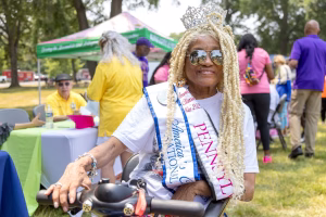 An elderly woman with blonde braids and a crown sits in a chair, surrounded by a crowd of people.