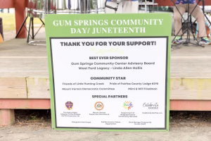 A green sign with white text on a wooden platform, likely at an outdoor event.
