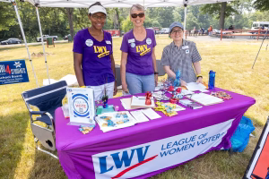 Three women volunteers for the League of Women Voters stand behind a table at an outdoor event.