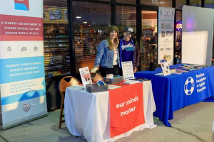Two women standing behind a table with a sign and a tablecloth