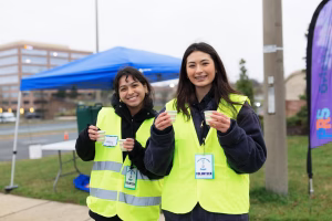 Two women in yellow vests standing outside, holding small cups, with a blue tent and a building in the background.