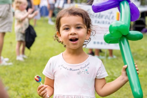 A happy young girl holding a lollipop and a balloon flower, enjoying an outdoor event.