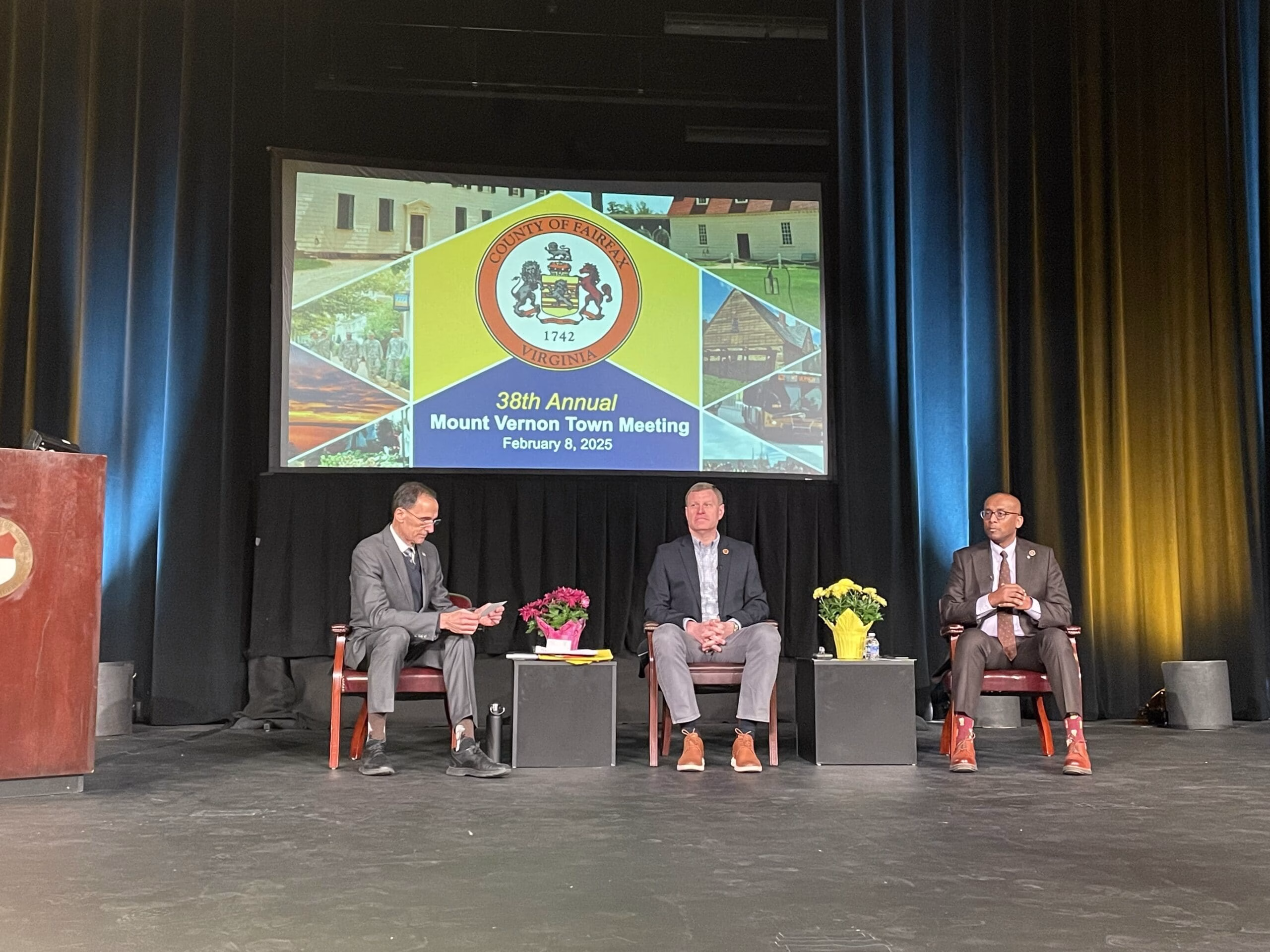 Four men in suits sitting on stage at the 38th Annual Mount Vernon Town Meeting, with a large screen behind them displaying the meeting's logo and date.