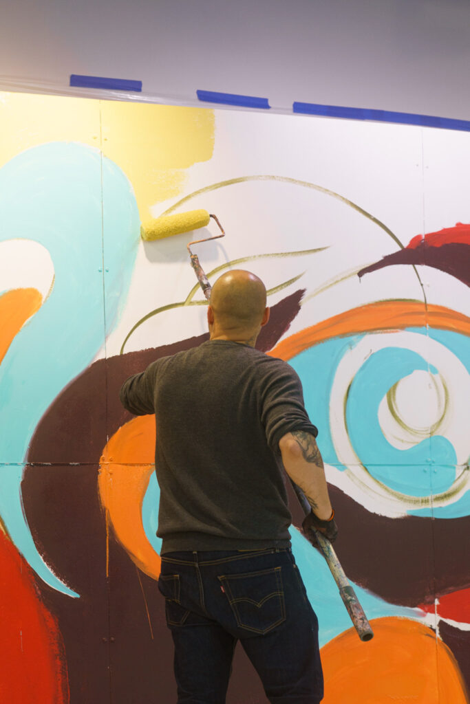 Rodrigo Pradel using paint roller to apply yellow paint to a top section of the indoor mural.